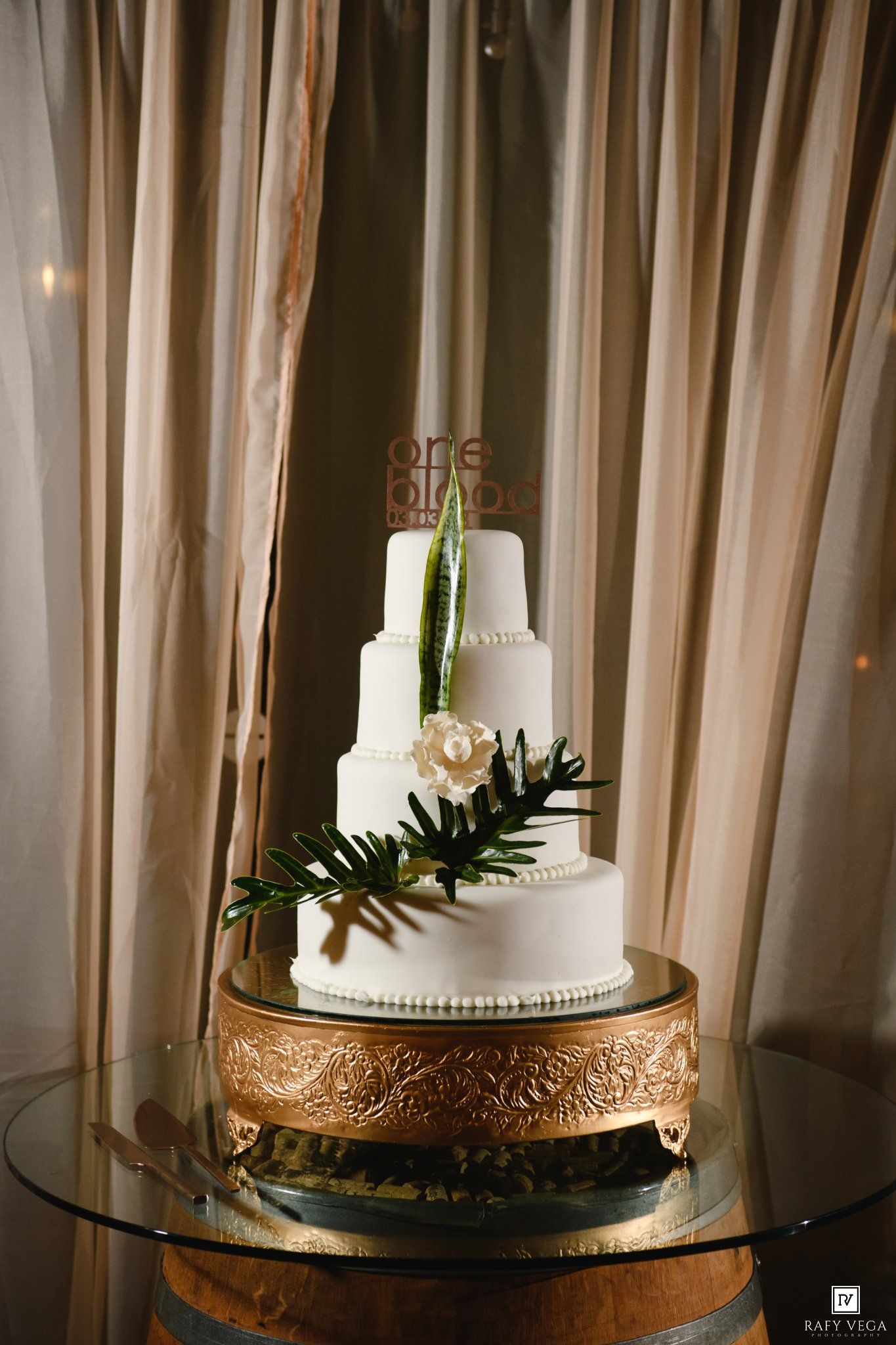 Elegant four-tier white wedding cake with greenery and floral decor on a gold stand, displayed against draped curtains.