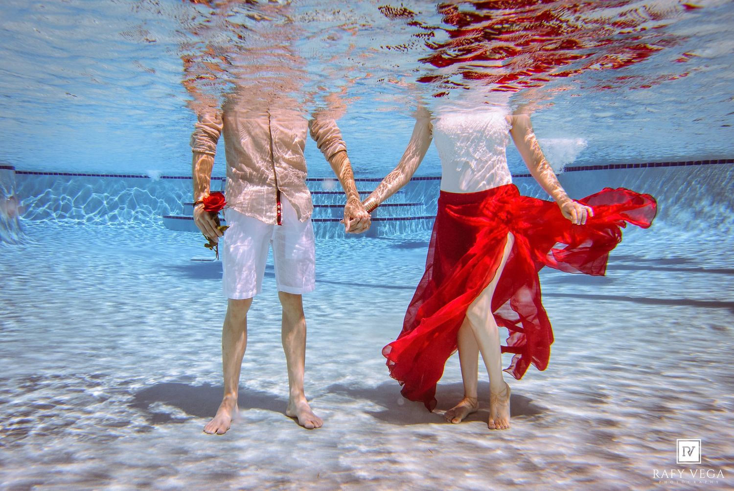 Couple holding hands underwater in a pool, woman in flowing red dress and man in white shirt, romantic underwater photography.