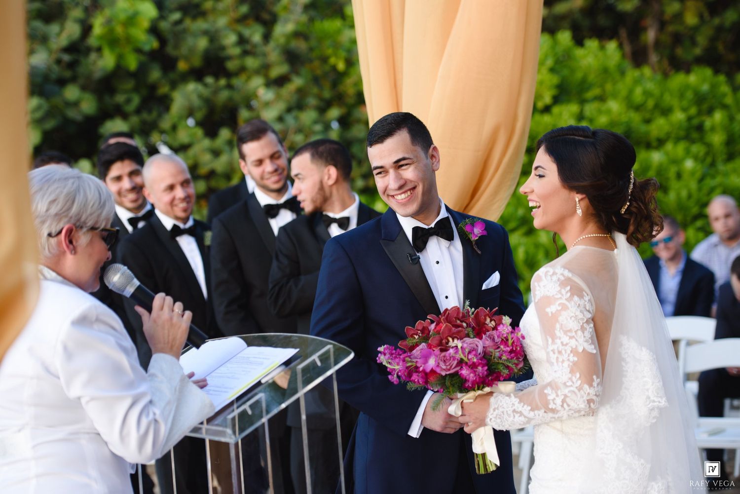 Smiling groom in tuxedo stands with bride holding pink bouquet during outdoor wedding ceremony with groomsmen.