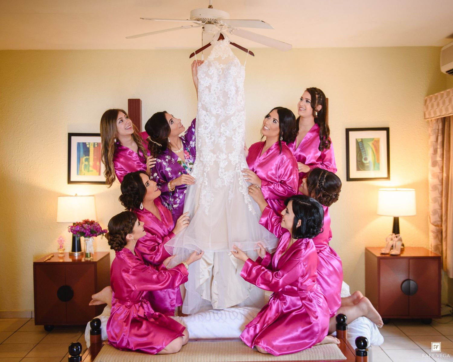 Bridesmaids in pink satin robes admiring a wedding gown hanging in a hotel room.