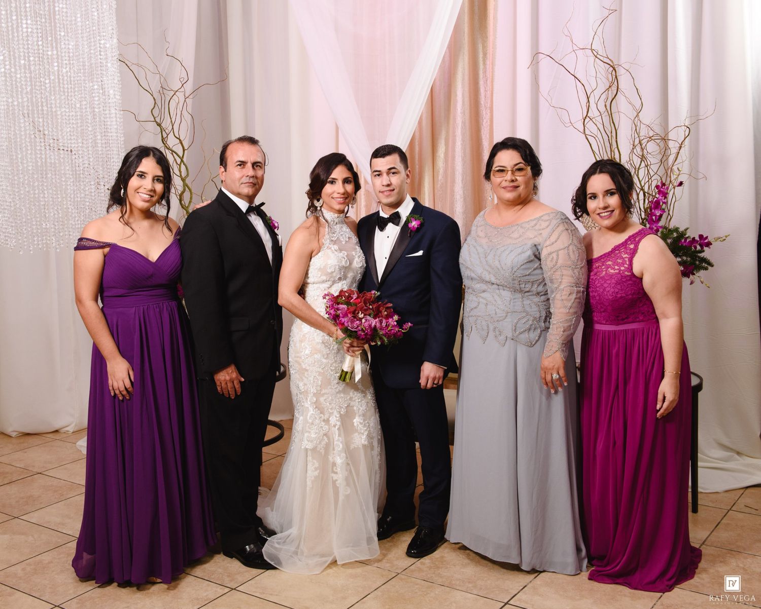 Wedding party posing together, bride in white gown with bouquet, groom in tuxedo, flanked by family in purple and gray formal attire.