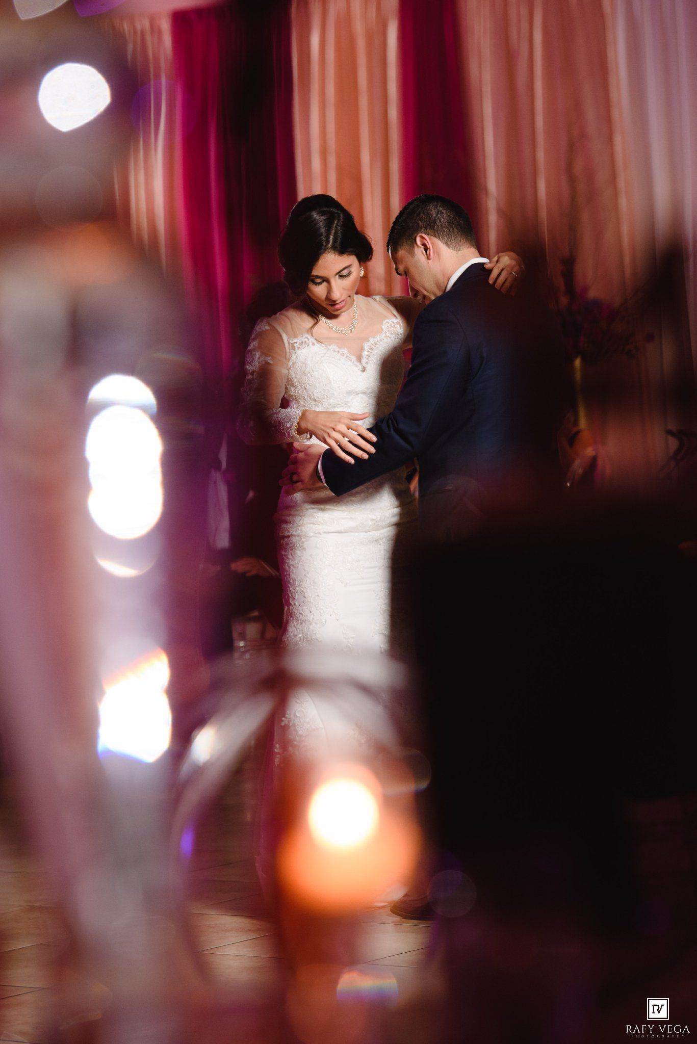 Bride and groom share an intimate moment during their first dance, framed by candlelight and pink drapes.