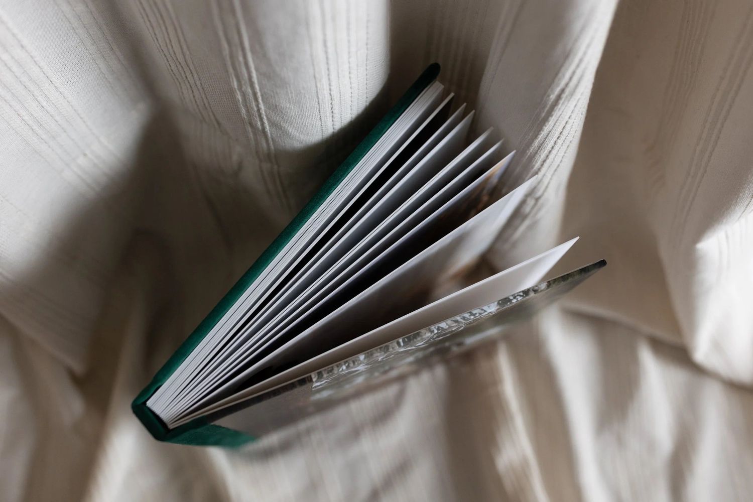 Open hardcover book with green spine lying on white fabric, pages fanned out showing thick layered sheets.