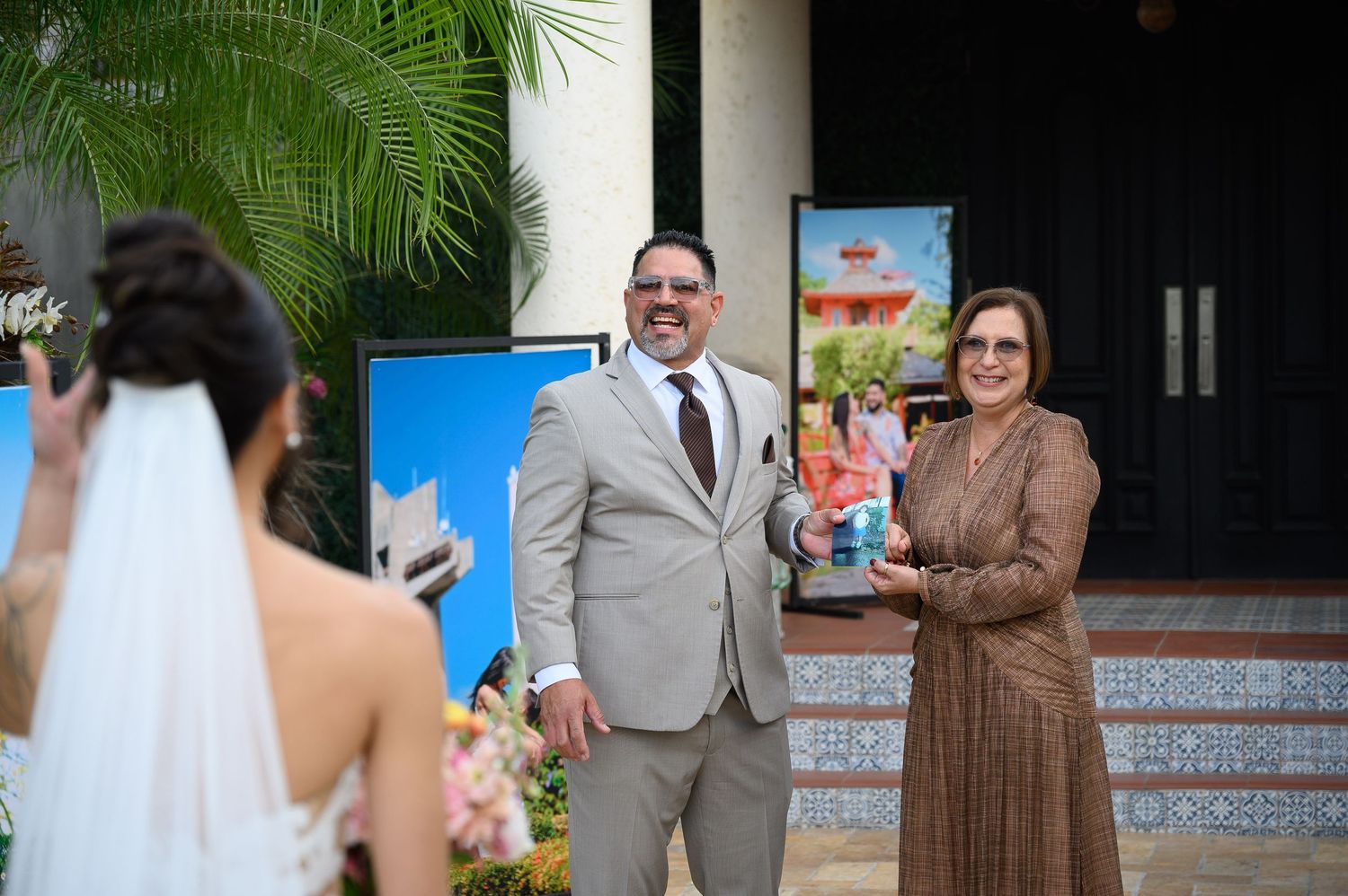 A bride in a white gown greets a smiling man in a gray suit and a woman in a brown dress at an outdoor wedding venue.