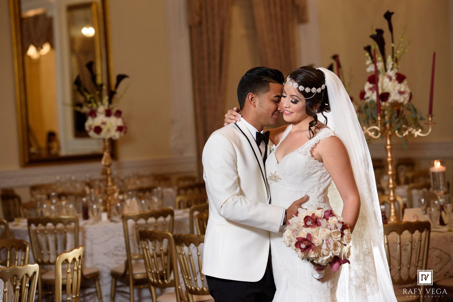 Bride and groom share a romantic embrace in an elegantly decorated wedding reception hall with gold chairs and floral centerpieces.
