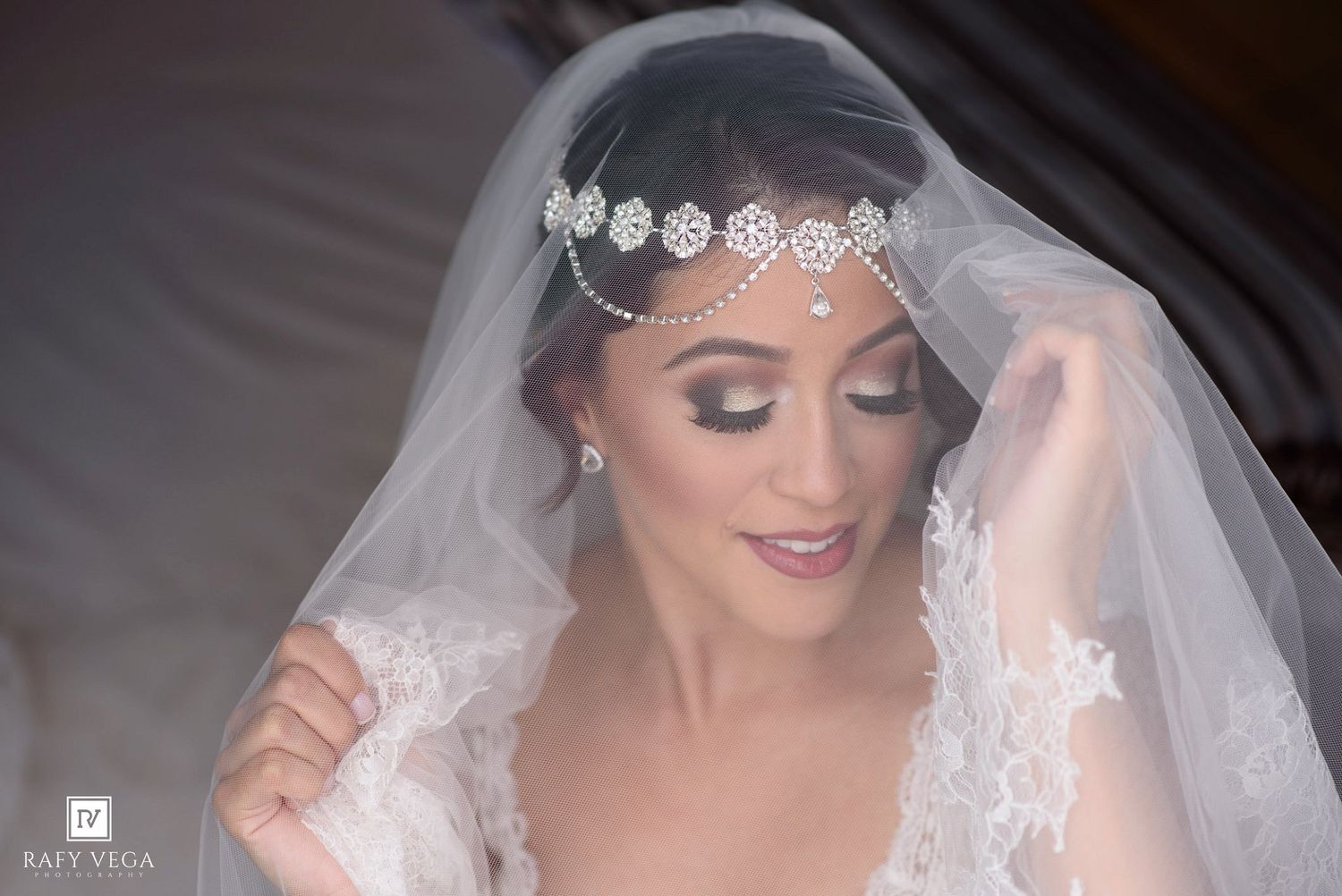 Bride in white lace gown wearing elegant crystal headpiece and veil with smoky eye makeup on wedding day.