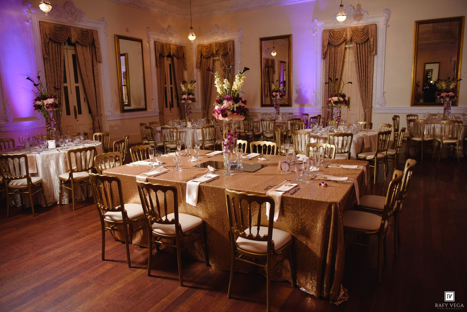 Elegant wedding reception hall with gold chiavari chairs, draped tables, floral centerpieces, and warm purple ambient lighting.