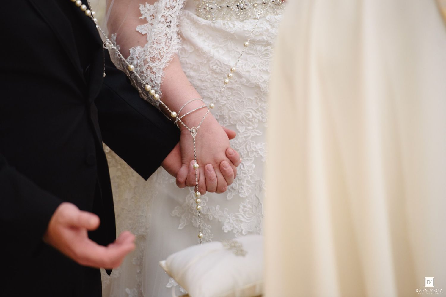 Bride and groom holding hands with pearl rosary at wedding ceremony, bride wearing lace gown.