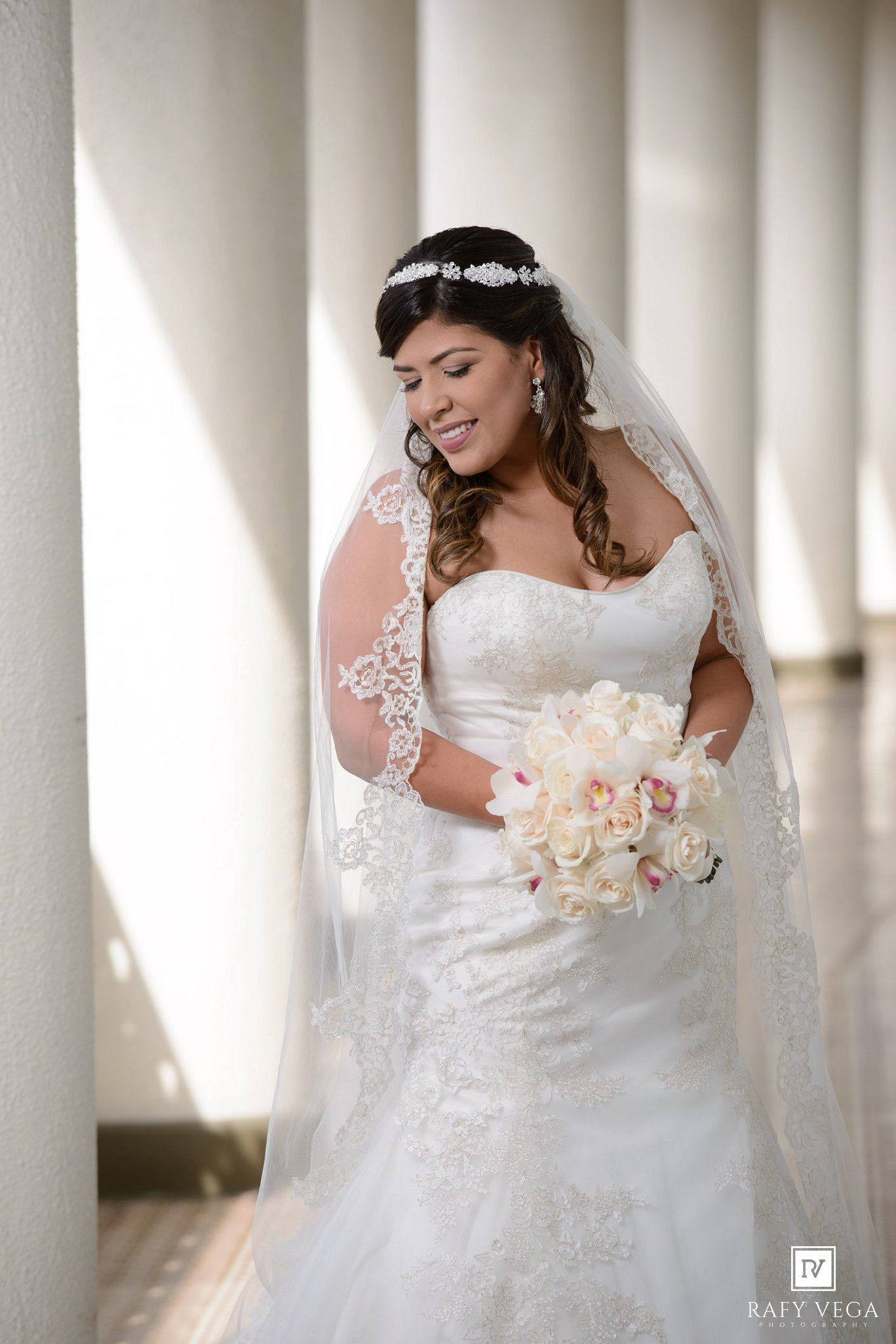 Bride in lace wedding gown and veil holding white bouquet, smiling among white columns.