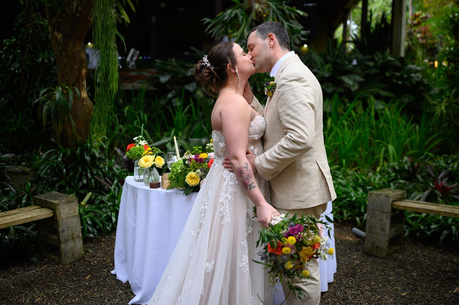 Bride and groom share a kiss outdoors surrounded by tropical greenery and colorful floral arrangements at their wedding.