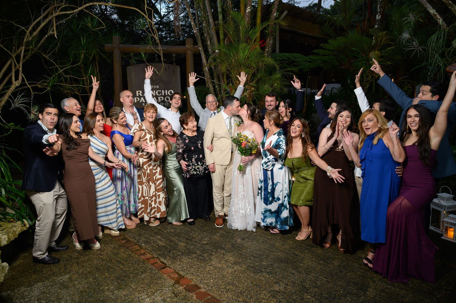 Large wedding group photo with guests celebrating outdoors at night, raising hands joyfully around bride and groom.