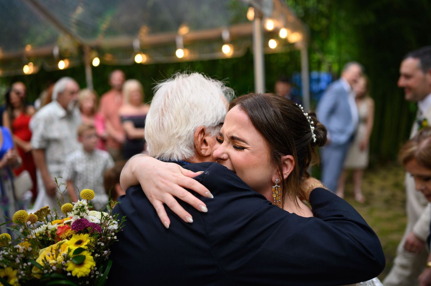 Bride in wedding dress hugging elderly guest at outdoor reception with string lights and sunflowers.