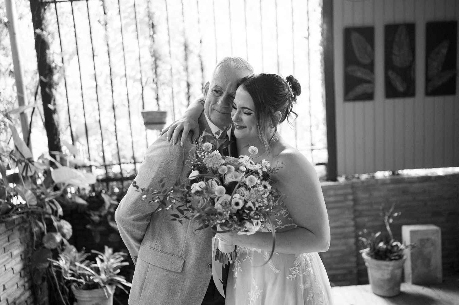 Black and white wedding photo of a bald groom embracing a smiling bride holding a floral bouquet outdoors.