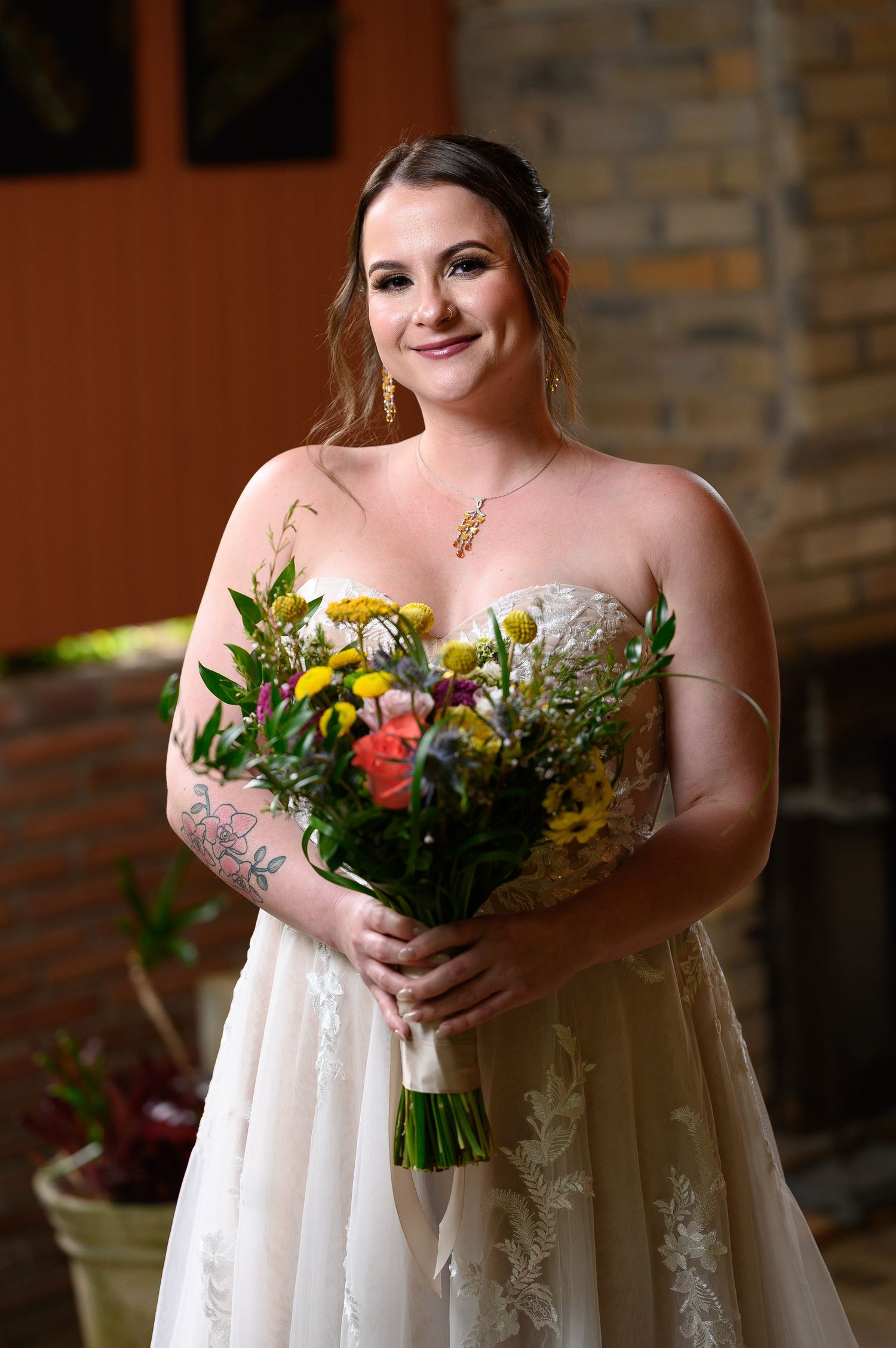Bride in strapless lace gown holding colorful wildflower bouquet with yellow, pink, and orange blooms, smiling outdoors.