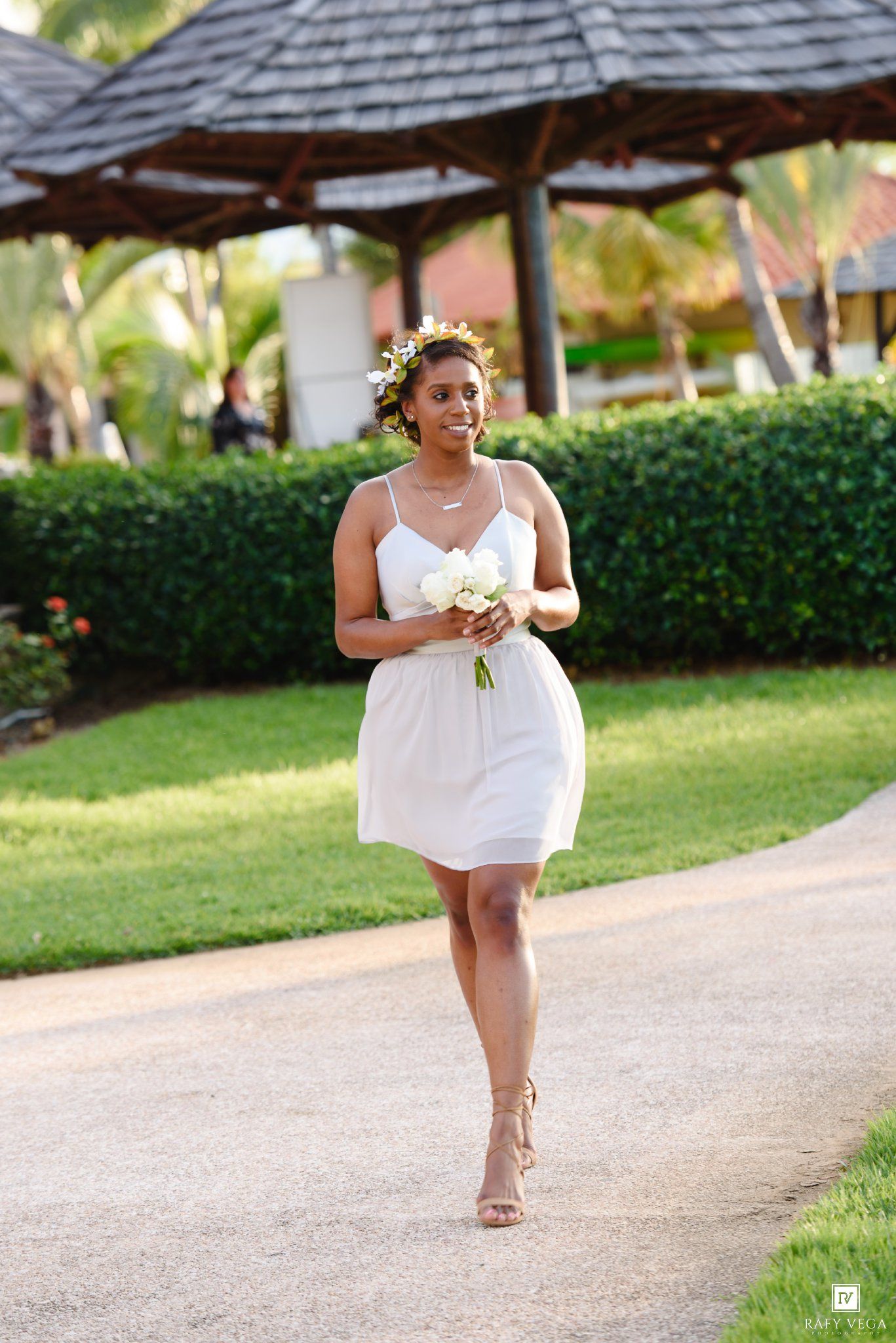 Smiling bride in white mini dress with floral crown carrying bouquet walking along garden pathway.