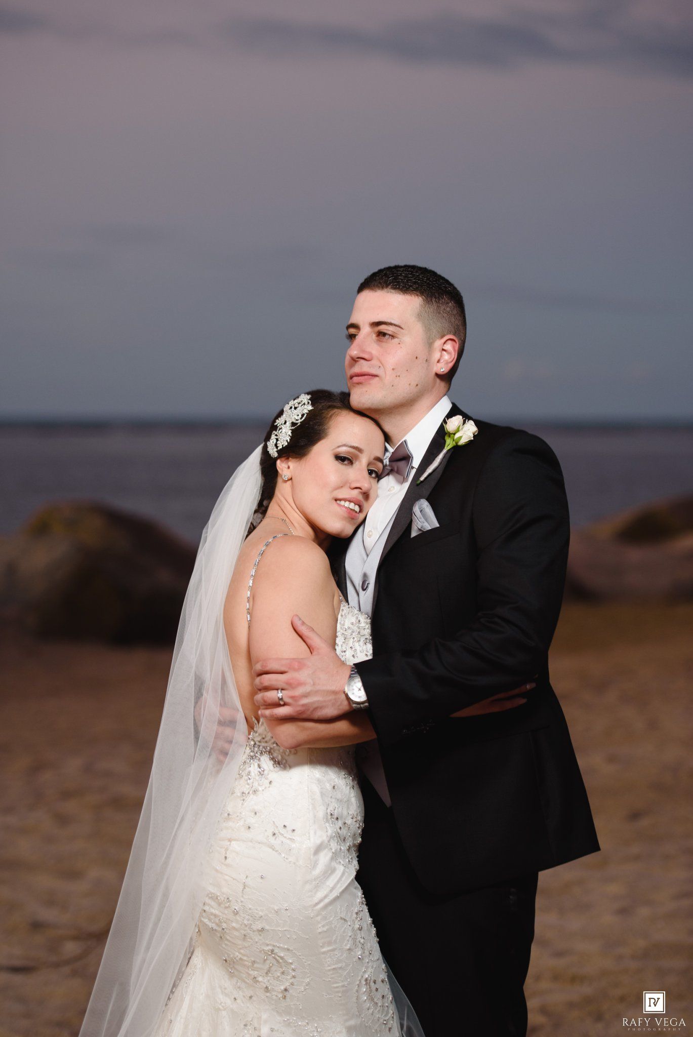 Bride and groom embrace on a beach at sunset, she in a beaded gown with veil, he in a black tuxedo.