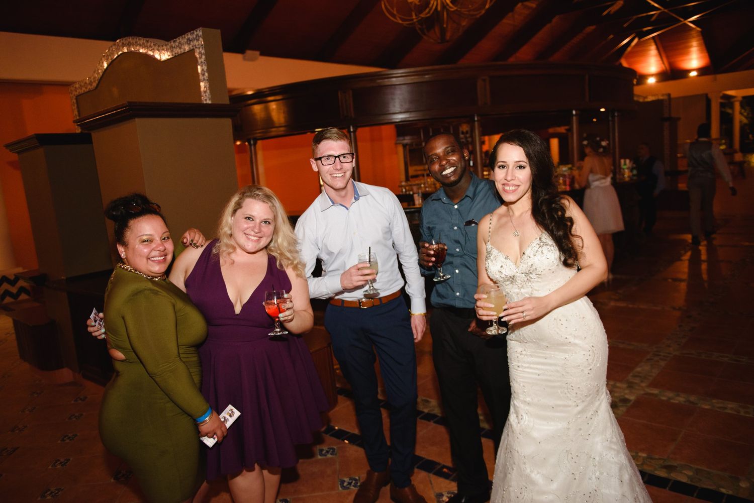 Bride in white lace gown poses with four guests holding drinks at an elegant indoor wedding reception venue.
