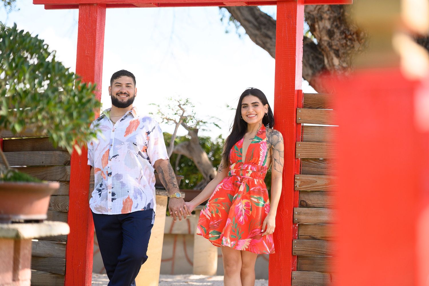 Couple holding hands under a red arch gateway, wearing floral outfits in a sunny garden setting.