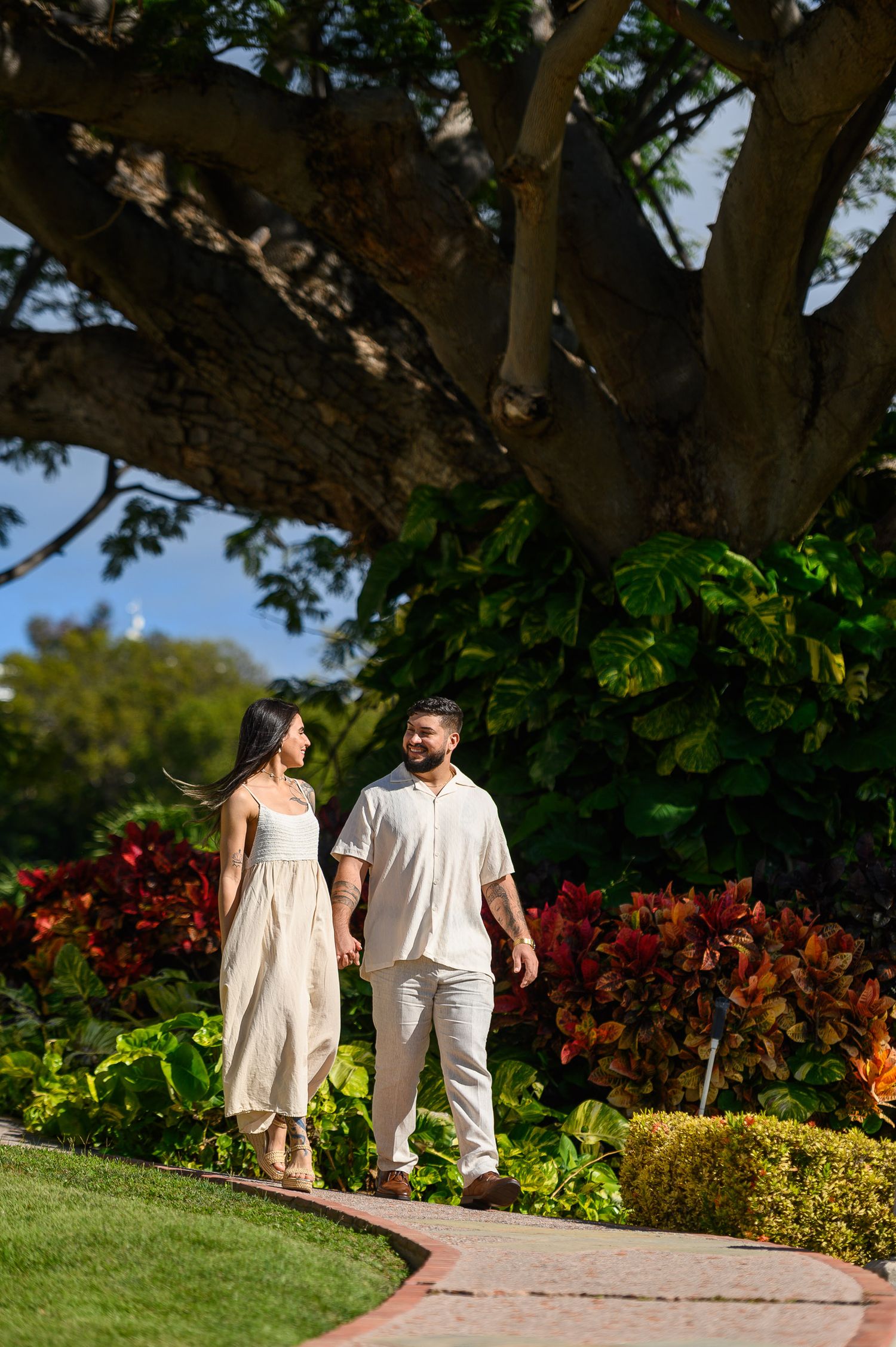 Couple in matching cream outfits walking hand in hand through lush tropical garden.