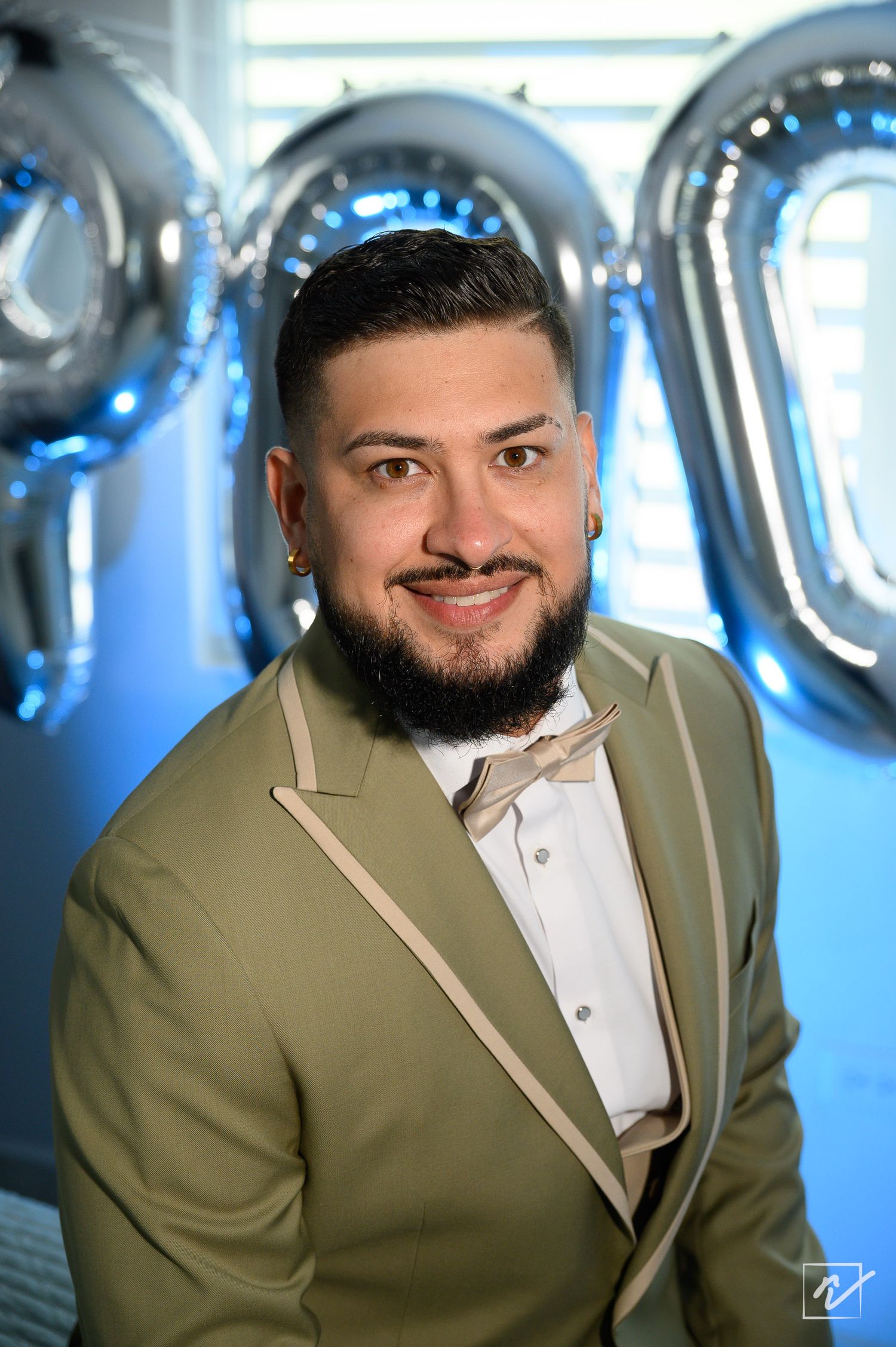 Smiling man with a groomed beard wearing an olive green blazer with cream trim and a beige bow tie, posed in front of silver balloons.