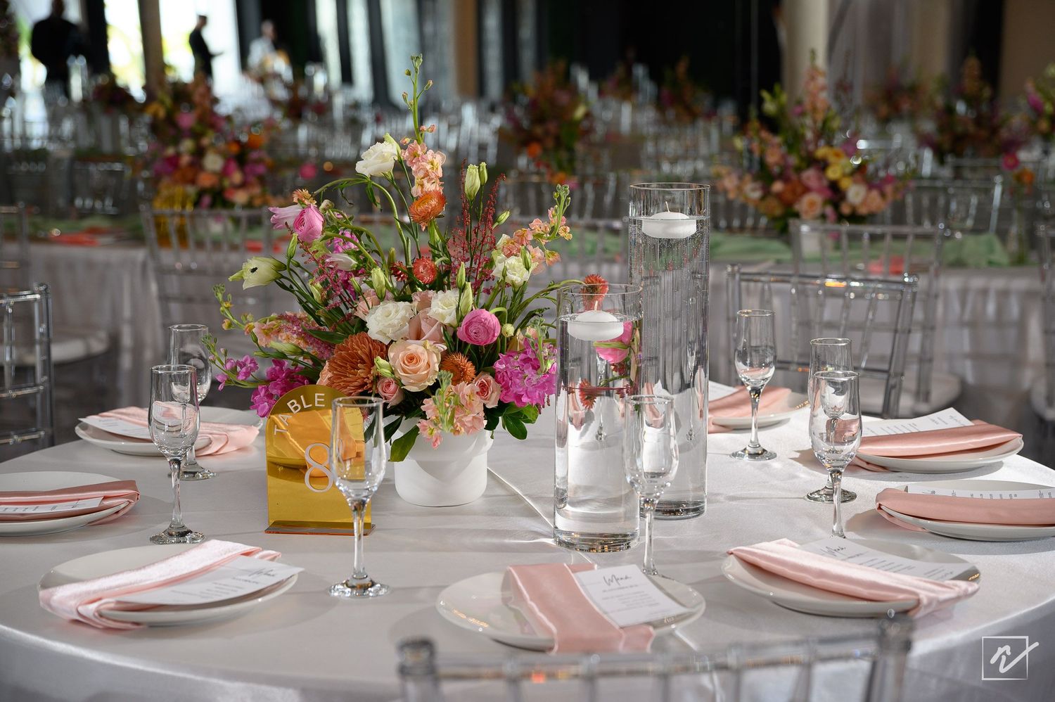 Elegant banquet table set with pink napkins, white plates, glassware and a colorful floral centerpiece of peach, pink and orange blooms.