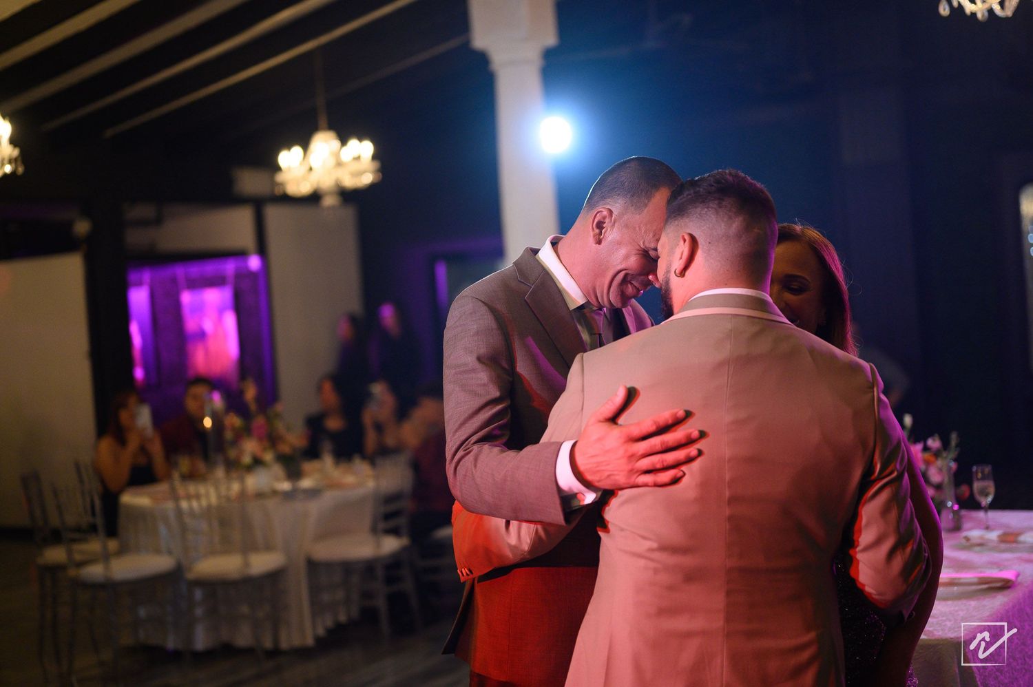 Two grooms in tan and brown suits share an intimate embrace during a first dance at an elegant wedding reception with guests seated nearby.