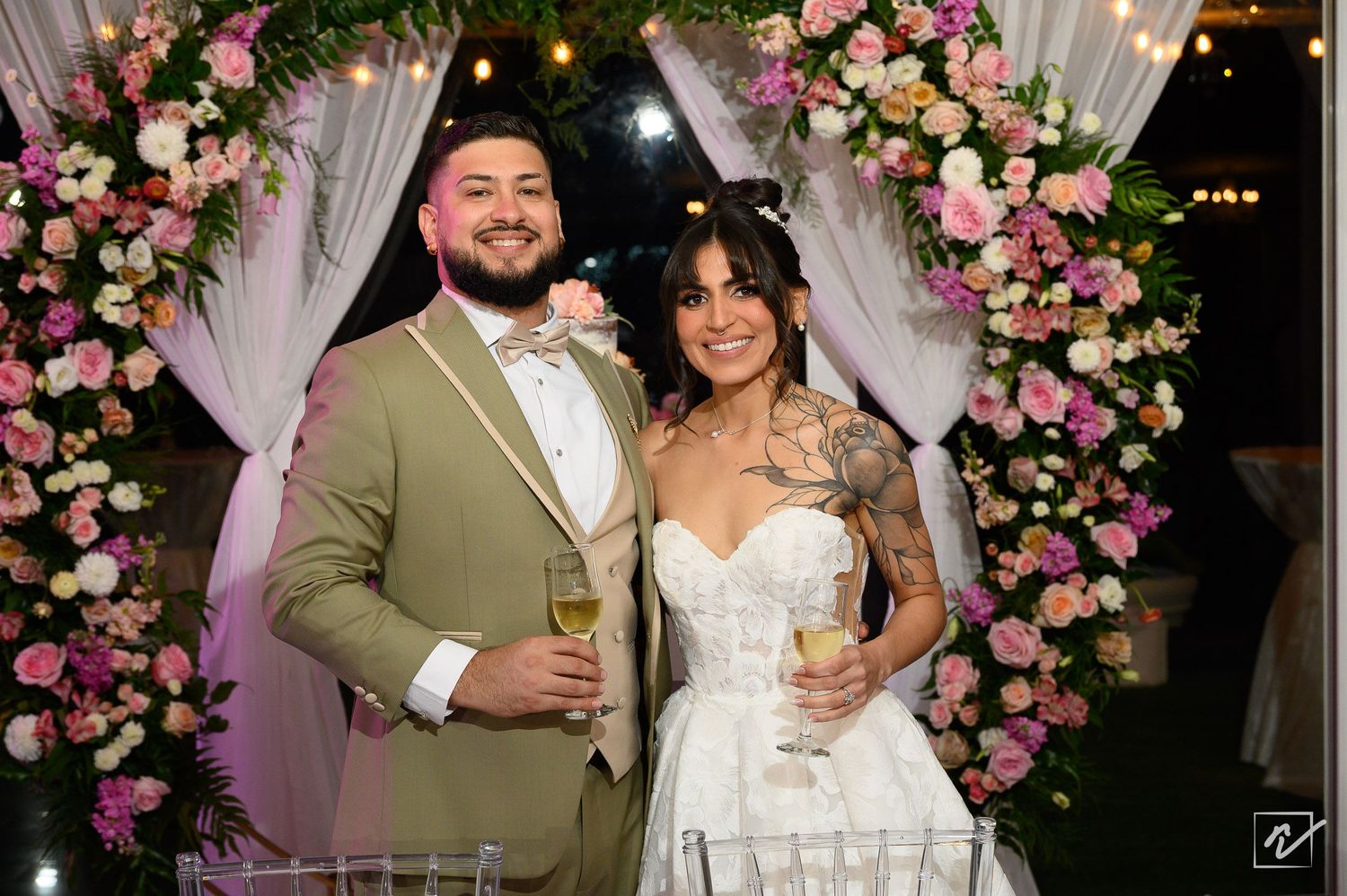 Smiling bride in a white strapless gown and groom in an olive suit hold champagne glasses before a floral arch with pink roses.