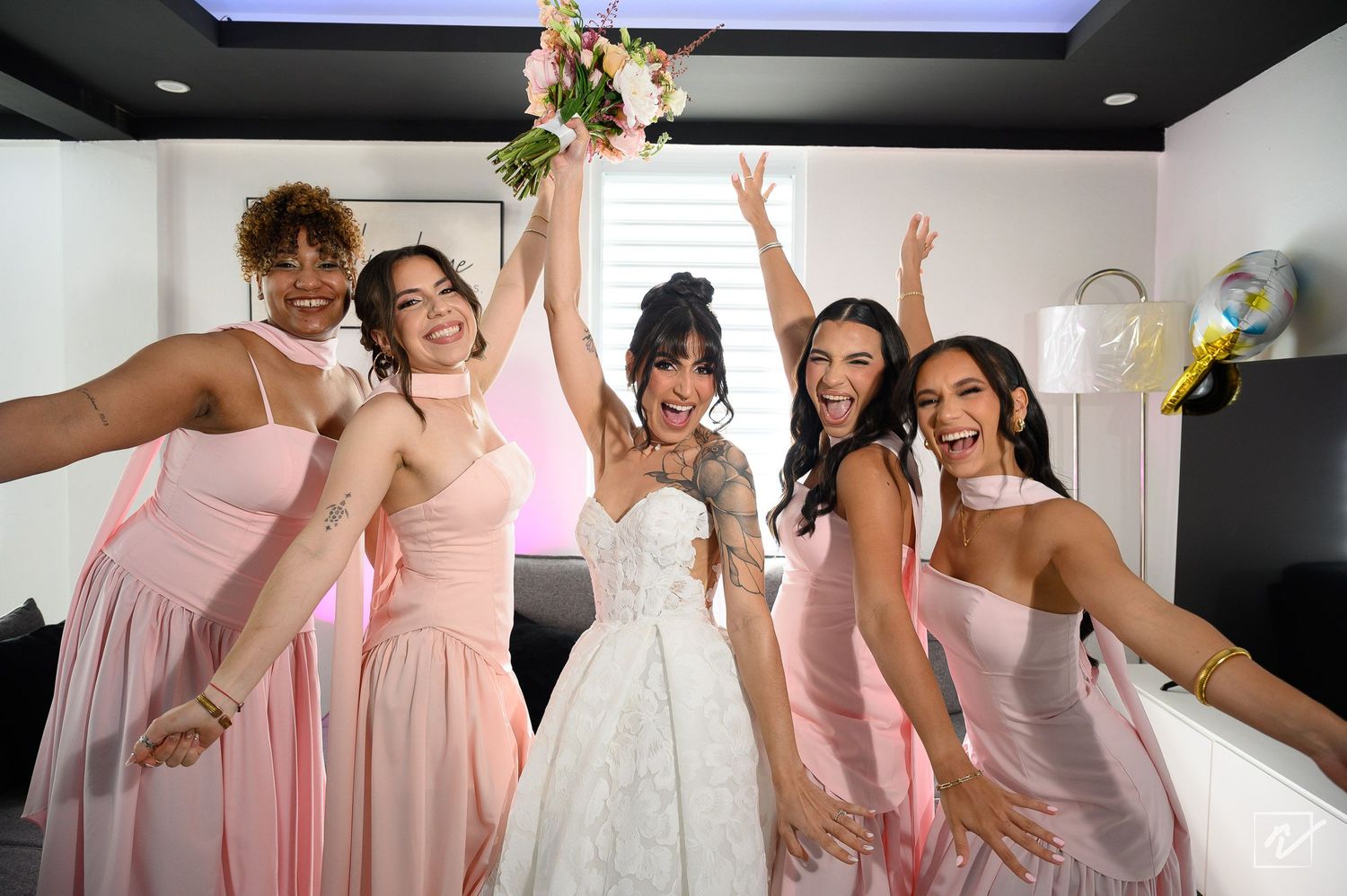 Bride in a white wedding gown holding a bouquet celebrates with five joyful bridesmaids in blush pink dresses.