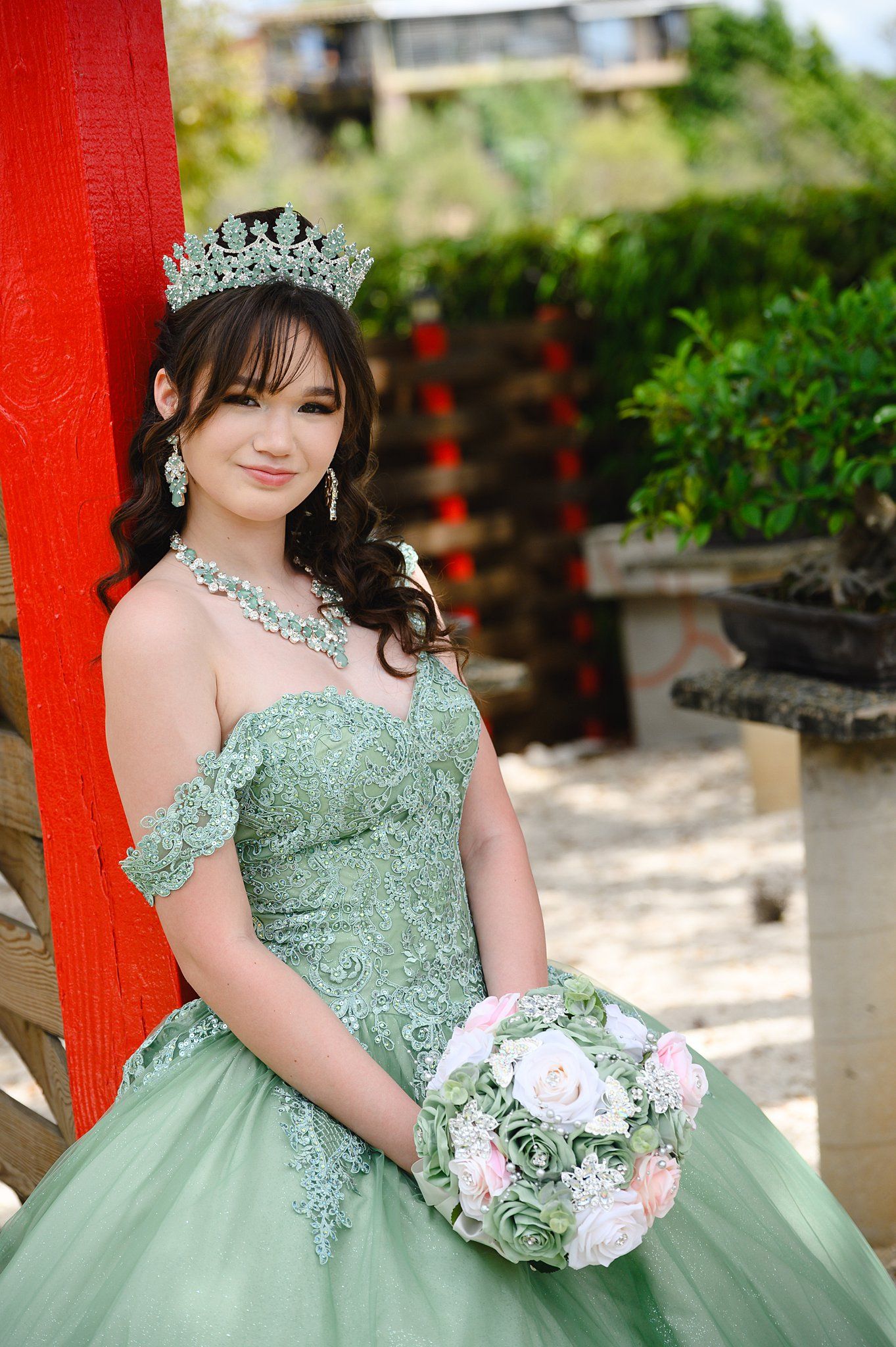 Young woman in sage green quinceañera gown with crystal crown holds floral bouquet outdoors.