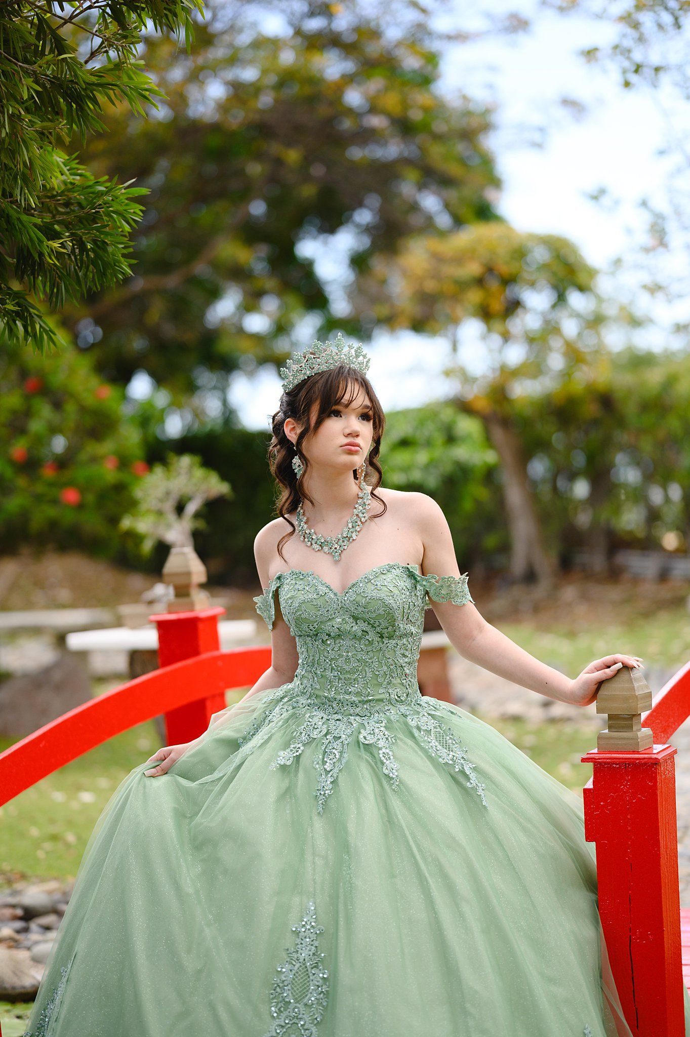 Young woman in sage green off-shoulder ball gown with tiara poses on red bridge surrounded by lush greenery.