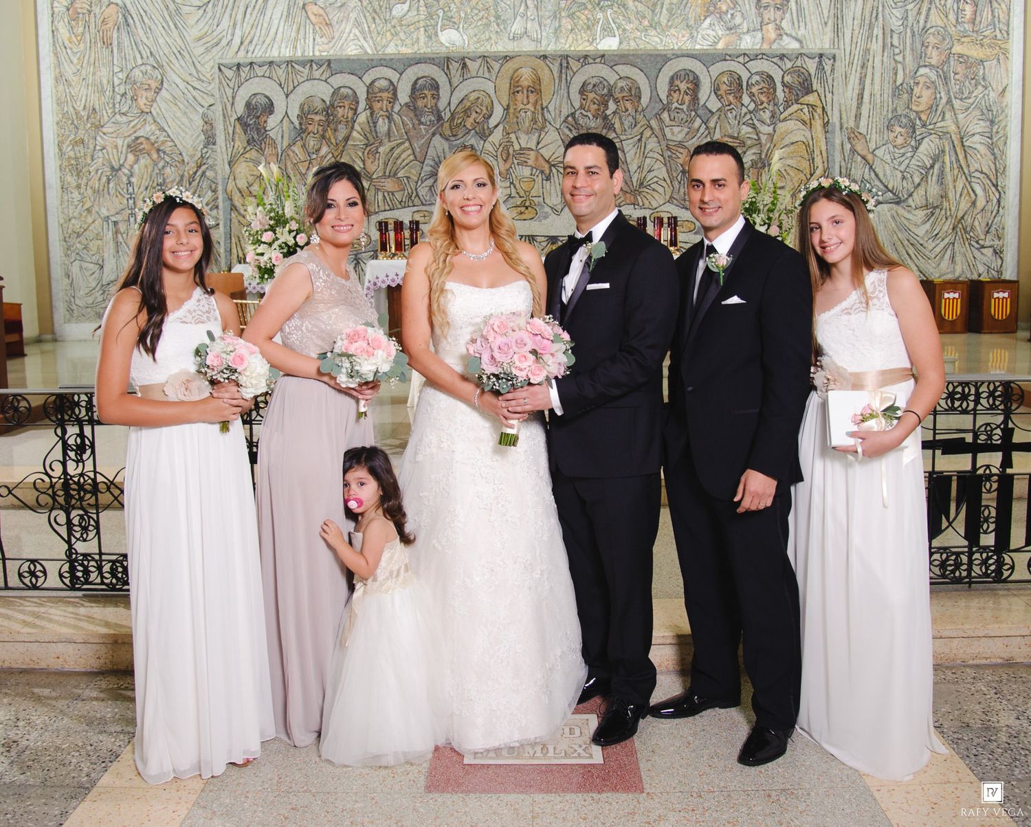 Wedding party poses in church with bride in white gown, groom in tuxedo, bridesmaids in blush gowns, and flower girl.