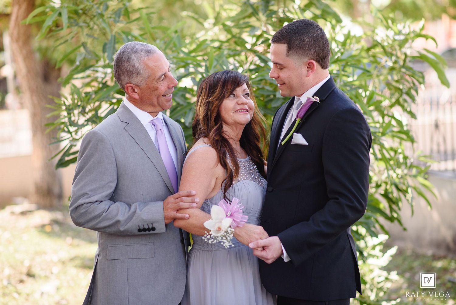 Family members smile together outdoors at a wedding, wearing formal attire with floral corsage and boutonnieres.