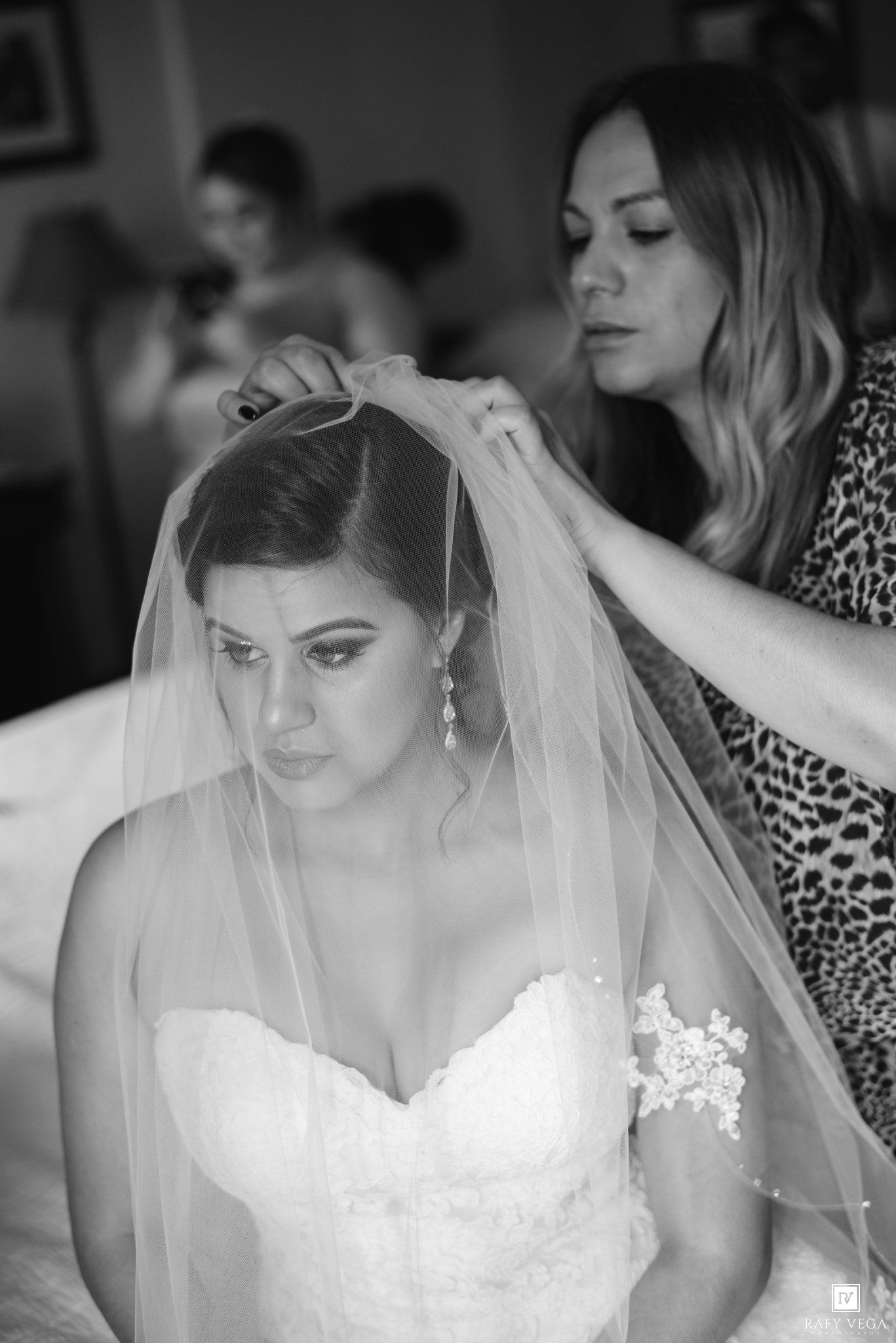 Black and white photo of a bride in a strapless lace gown having her veil adjusted by a woman in a leopard print top.