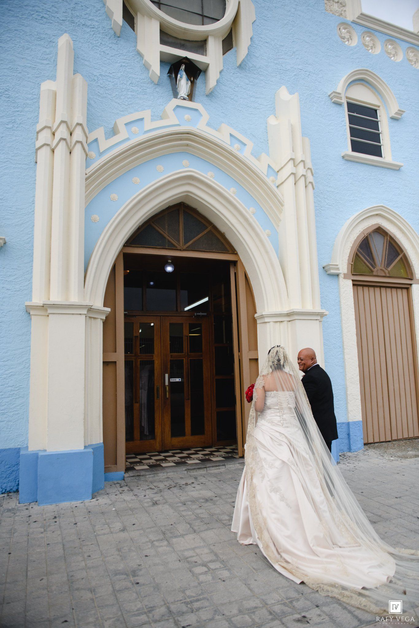 Bride in white gown and groom entering a blue church with ornate white arched doorway on wedding day.