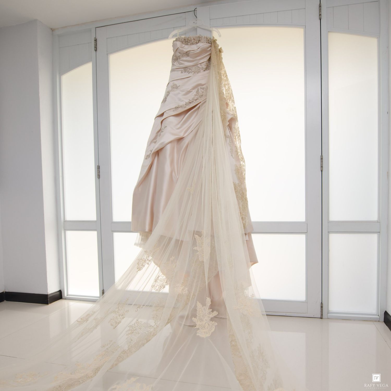 Elegant blush pink wedding gown with lace details and long train hanging on display near bright windows.