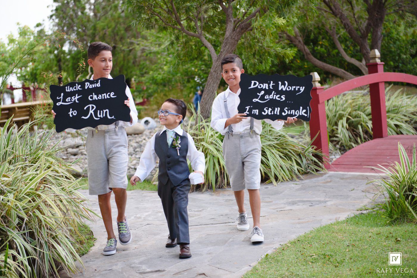Three boys at a wedding holding chalkboard signs with humorous messages, dressed in formal attire outdoors.