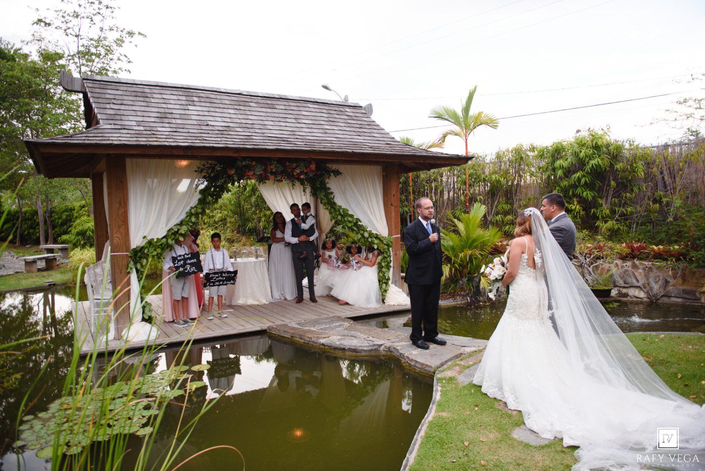 Outdoor wedding ceremony at a tropical garden gazebo with a pond, bride and groom standing before an officiant.