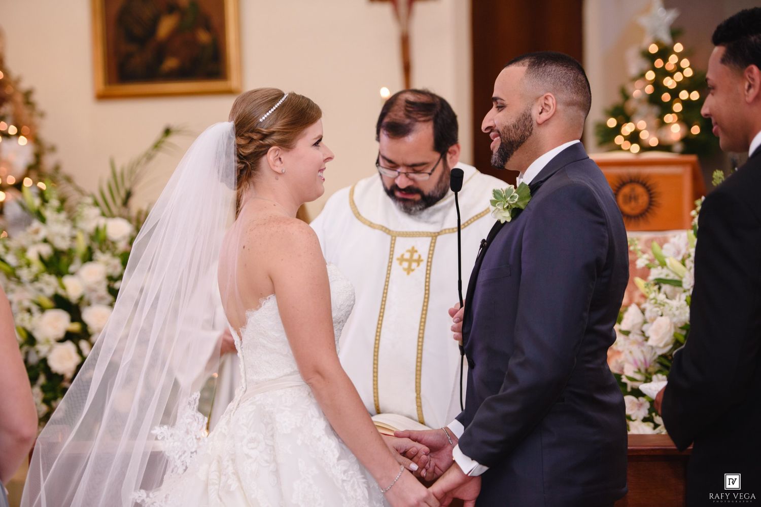 Bride and groom hold hands before priest during romantic church wedding ceremony.