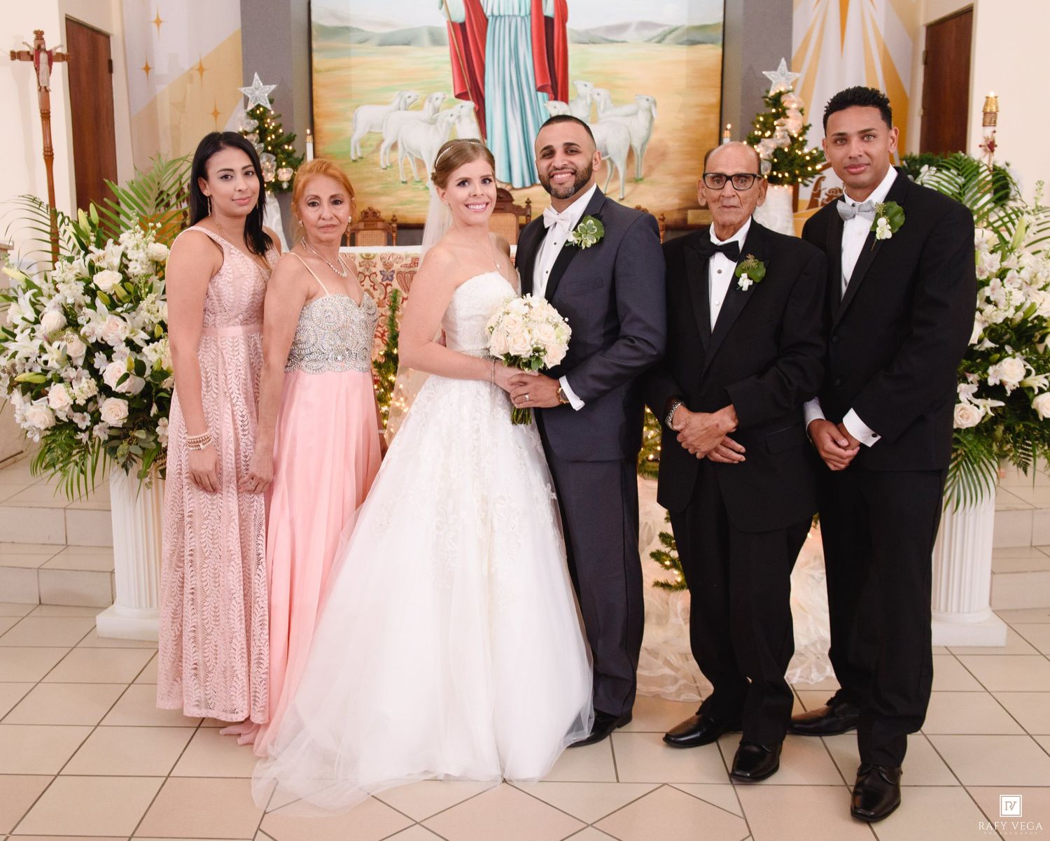 Wedding party poses with bride and groom at church altar surrounded by white floral arrangements.