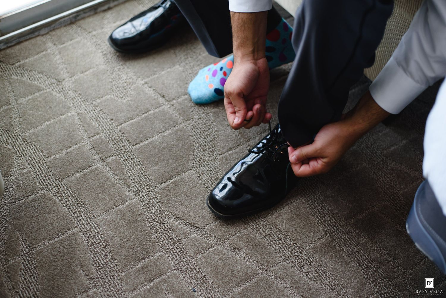Groom tying black dress shoes on carpet floor, with colorful socks visible, preparing for wedding day.
