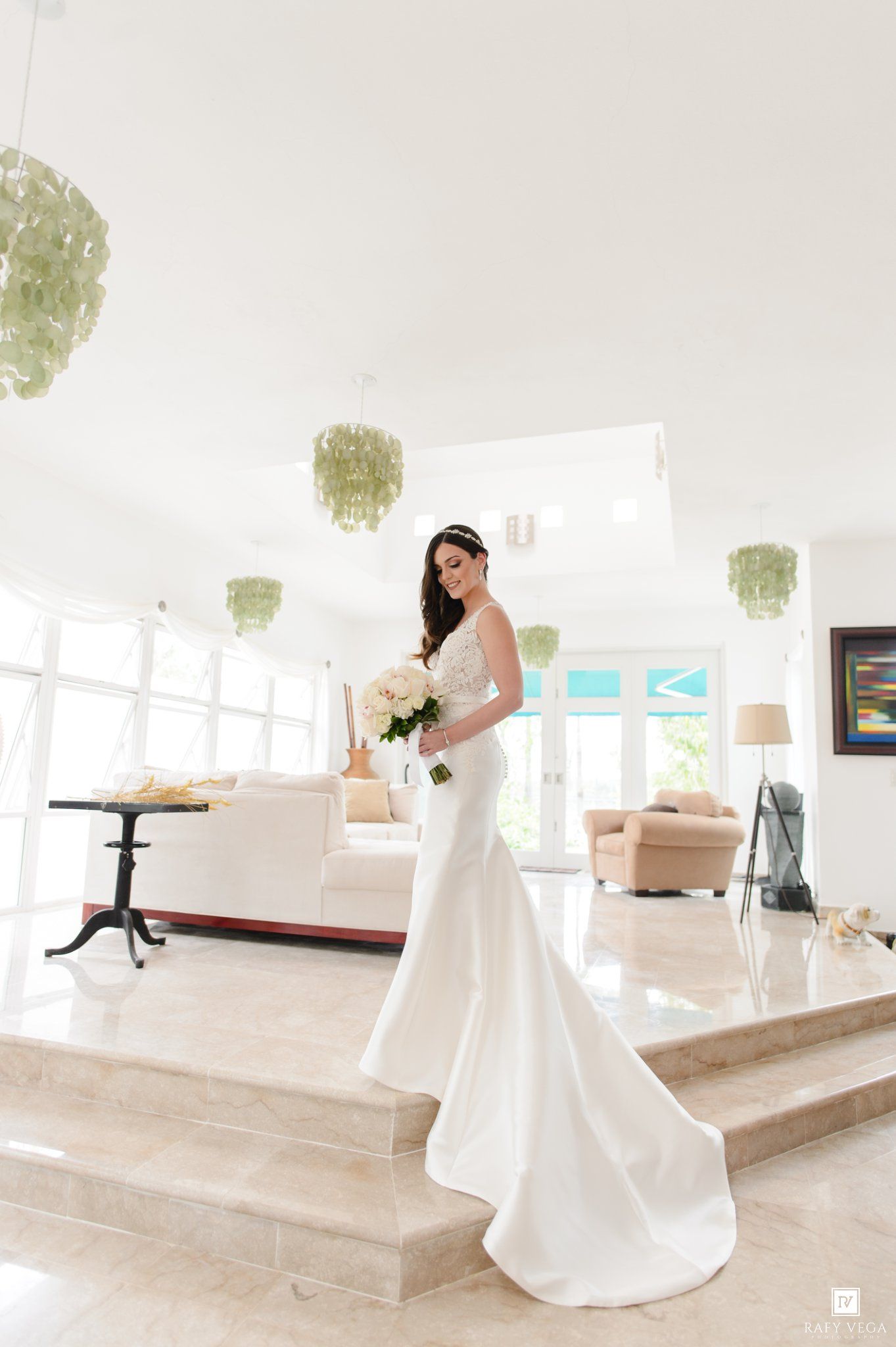 Bride in white mermaid gown holding bouquet stands on marble stairs in elegant bright venue with green chandeliers.