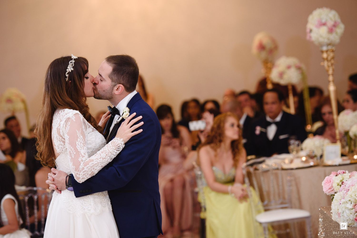Bride and groom share first kiss at elegant wedding reception with guests watching in background.