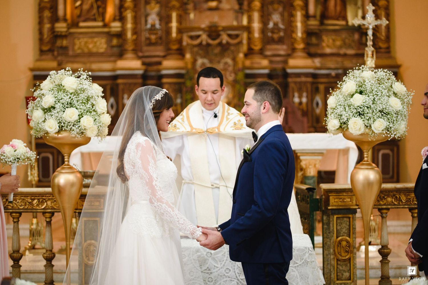 Bride and groom exchange vows at elegant church altar with white floral arrangements and officiant.