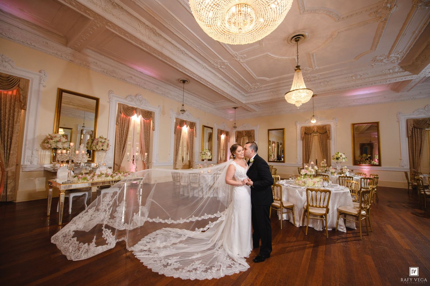 Bride and groom share a romantic moment in an elegant ballroom with chandelier, gold chairs, and a flowing lace wedding veil.