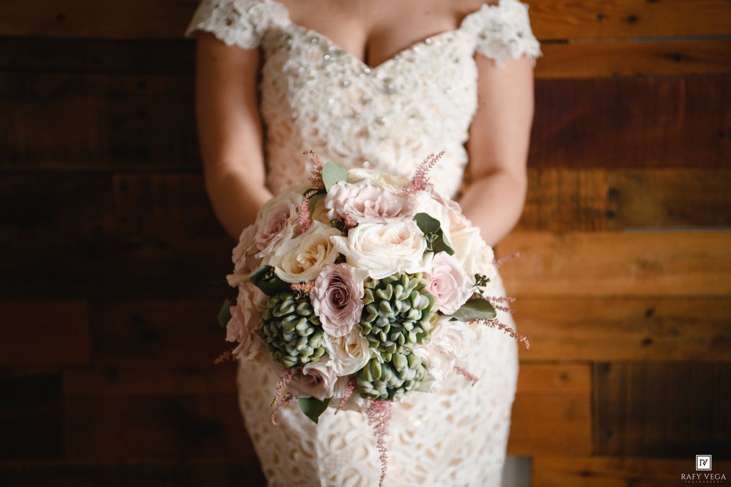 Bride in lace wedding gown holding elegant blush pink and green bouquet with roses and hydrangeas against rustic wood wall.