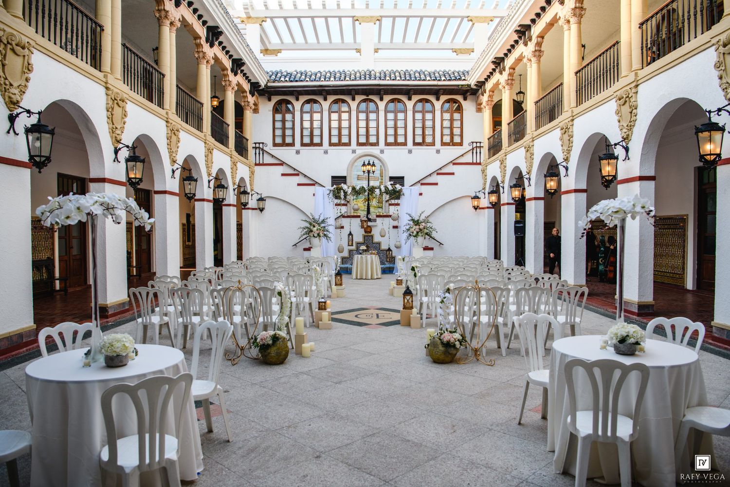 Elegant outdoor wedding ceremony setup in a historic Spanish courtyard with white chairs, floral arrangements, and arched architecture.