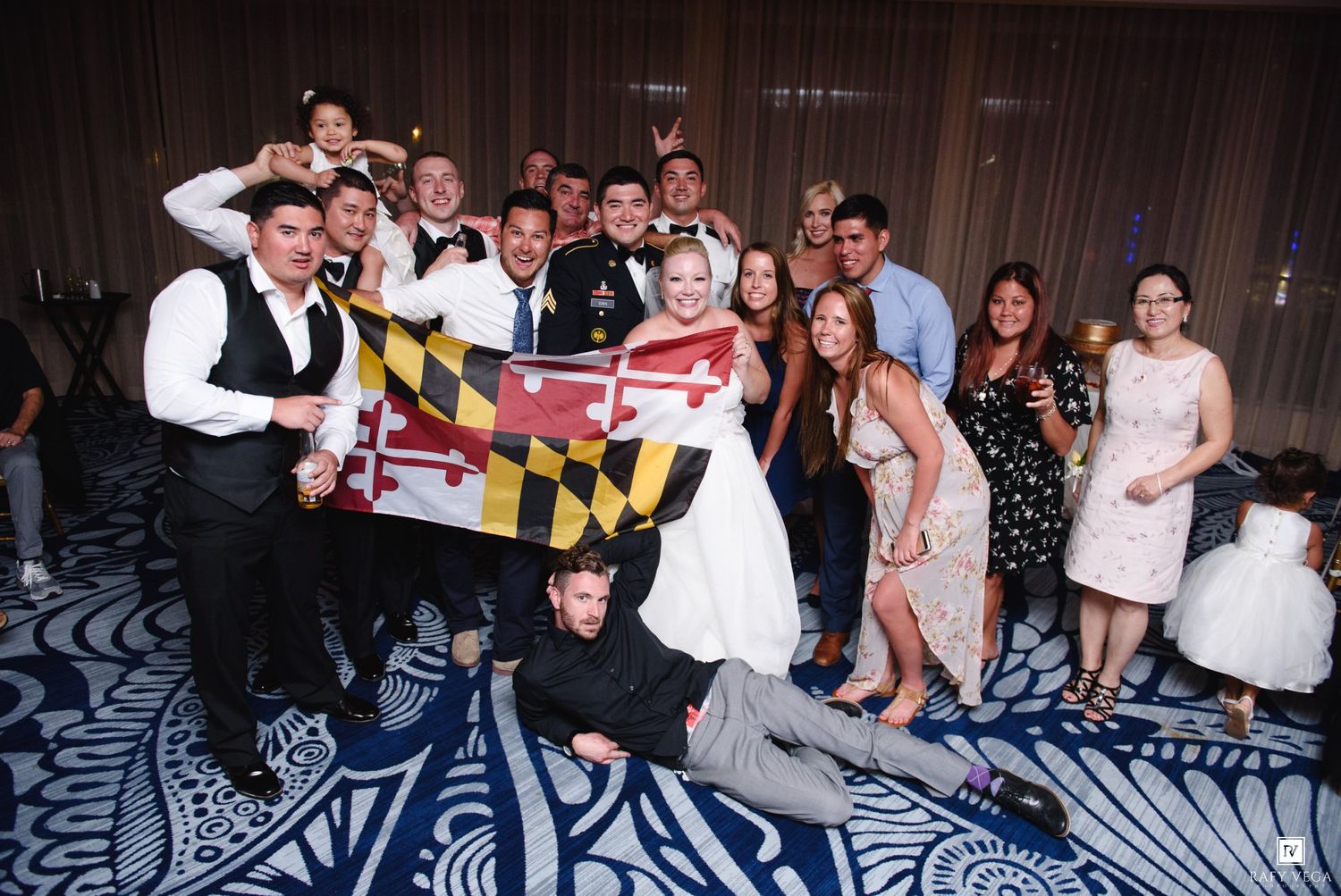 Large wedding party group photo with guests holding a Maryland state flag at the reception.