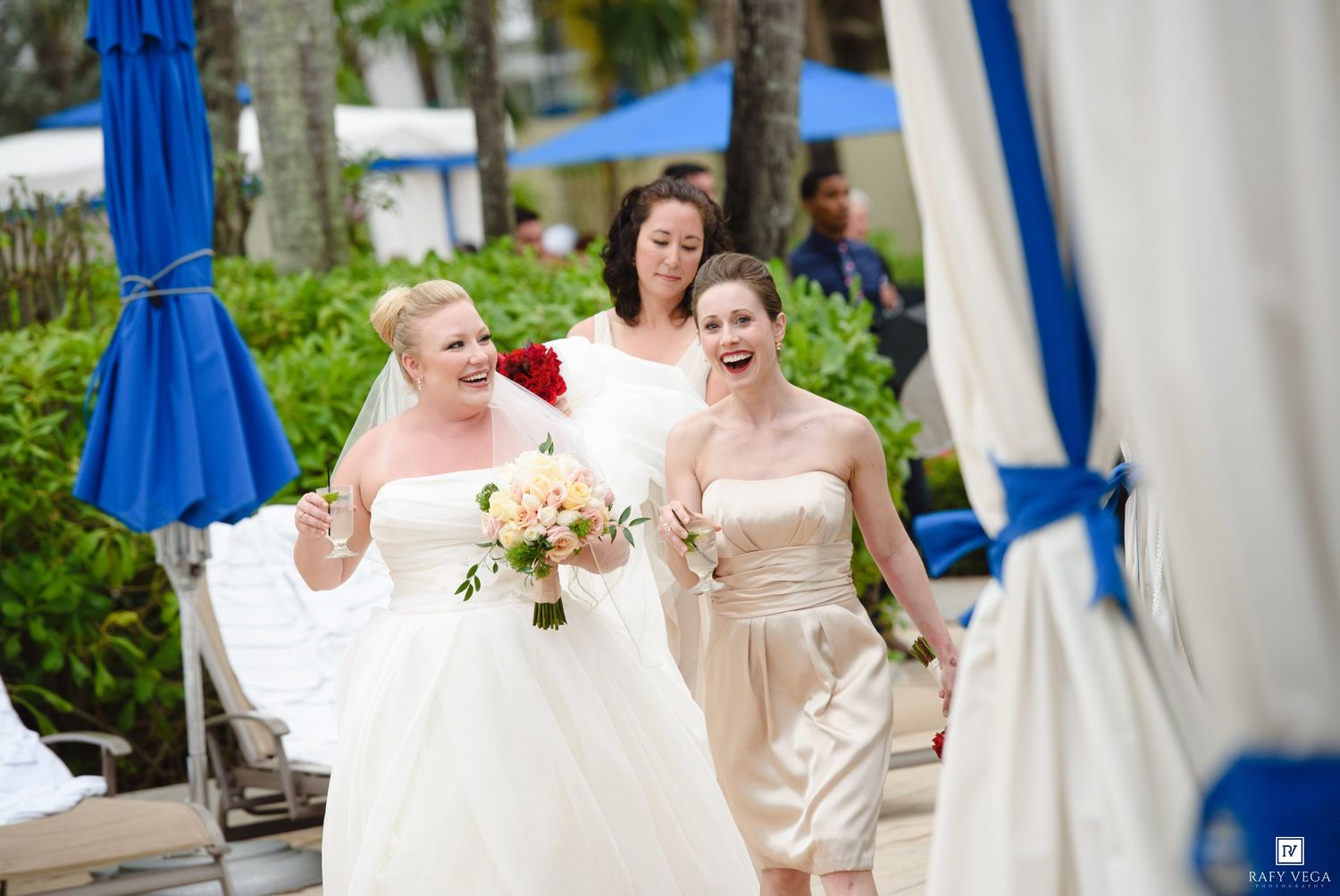 Bride in white gown walks with bridesmaids in champagne dresses near a poolside venue with blue umbrellas.