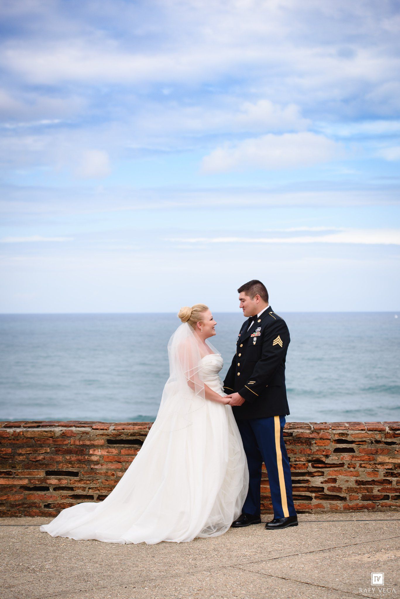 Military groom in dress uniform holds hands with bride in white gown at ocean overlook with brick wall.