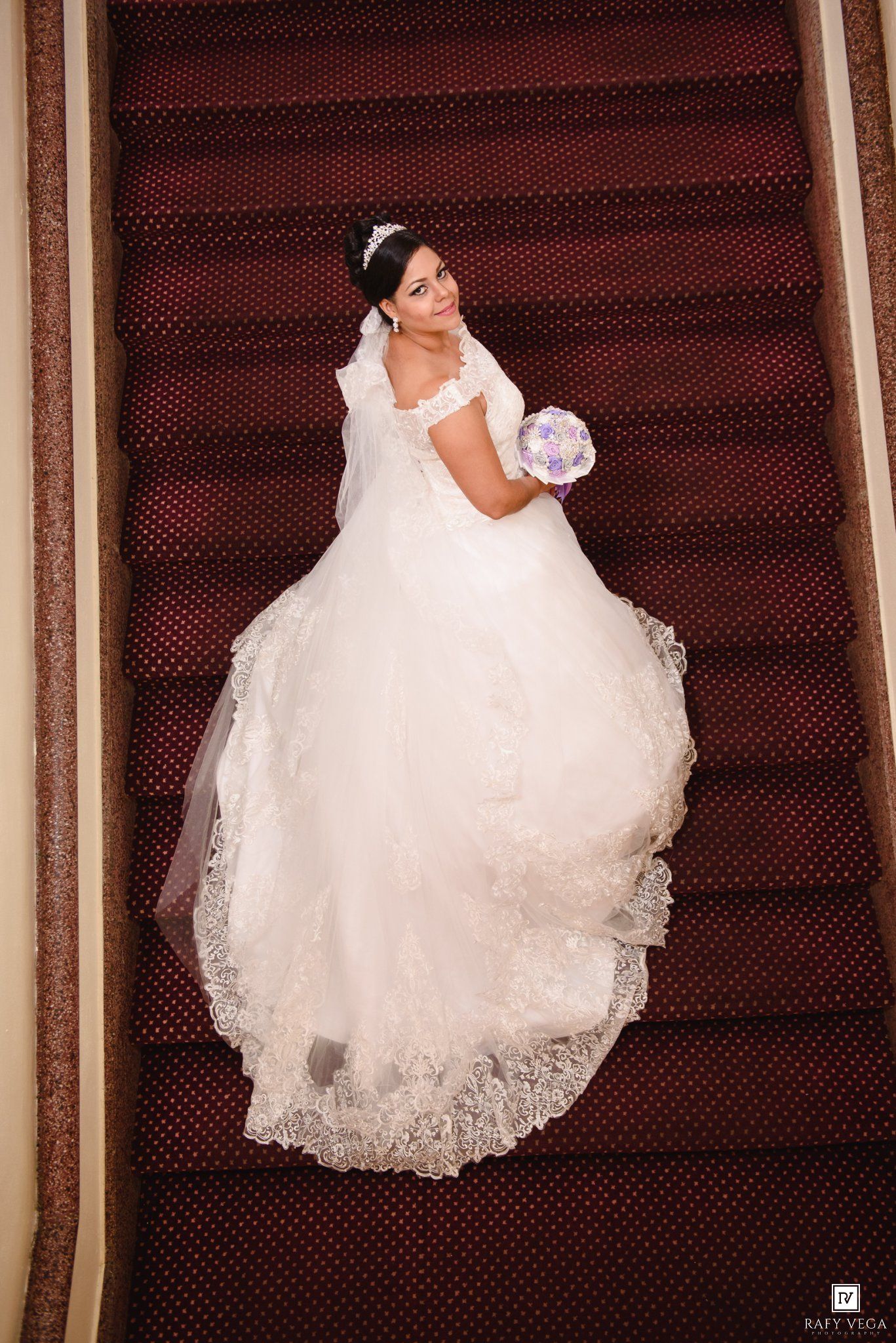 Bride in elegant white ball gown with lace trim holds bouquet on stairs, photographed from above.